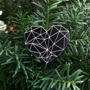 Black heart-shaped reflector badge with geometric white pattern, displayed against a vibrant green tree background